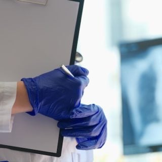 A pleural mesothelioma trial doctor holds a clipboard showing 100% survival of study patients one year after treatment. The clipboard and the doctor's gloved hands are in the foreground on the left, and an out-of-focus chest x-ray is in the background on the right.