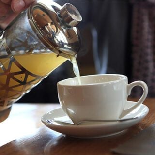 A photo showing yellowish-colored tea being poured from a glass and metal teapot into a white teacup.
